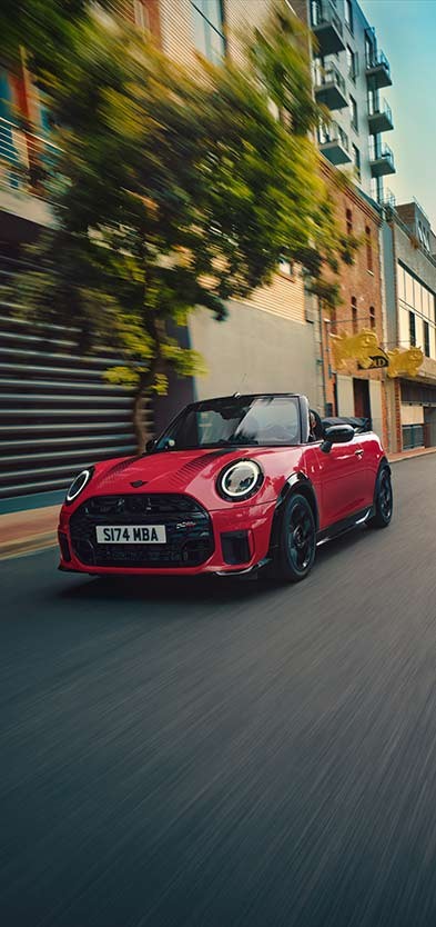 Frontansicht eines MINI Cooper Cabrio in Copper Grey an einem Strand mit zwei daneben stehenden Personen.